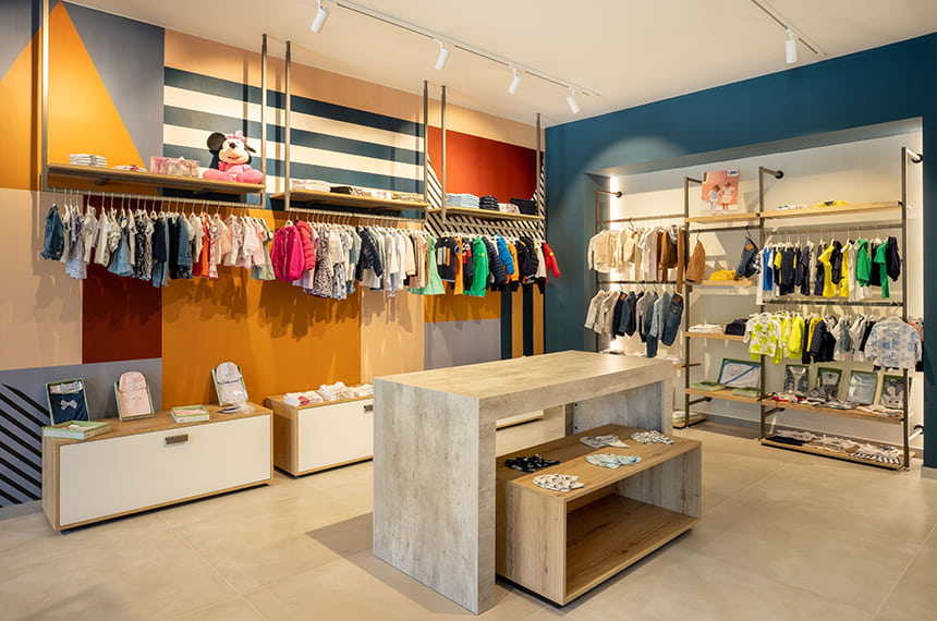 Vista parete espositiva negozio abbigliamento giovane, zona kids con parete espositiva e tavoli di vendita. Spettacolo design per Rolema Noi 2 Young
